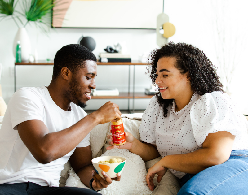 Two young people share a drink and a bowl of fruit on a white couch in a nice home.