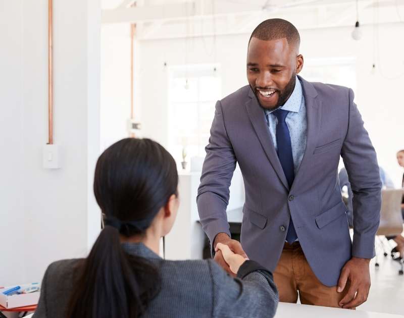 man wearing a suit smiling and shaking a woman's hand as he enters for a job interview
